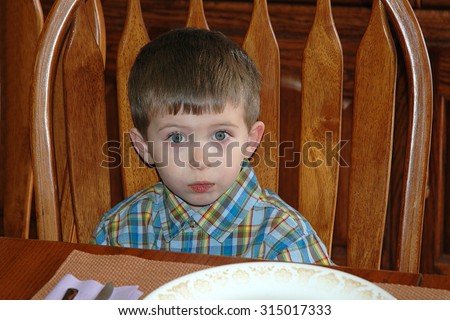 Young caucasian preschool age boy sitting at a wood dining room table waiting for food. Not sure he wants what is being served. concerned expression.
