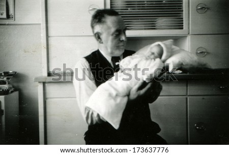 GERMANY - 1950s: An antique photo shows man in a waistcoat, white shirt and tie holding a baby in diapers, sitting in the kitchen