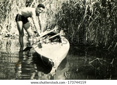GERMANY - 1950s: An antique photo shows man standing ankle-deep in the water near the paddle boats on a background of reeds