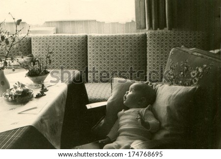 GERMANY -  CIRCA 1950s: An antique photo shows baby lying on pillows on a sofa near the table with a bouquet of flowers and looks aside window