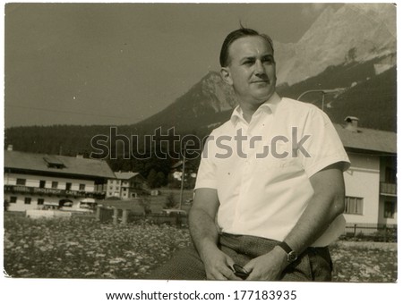 GERMANY - CIRCA August, 1965: An antique photo of middle-aged man in a white shirt with short sleeves posing against mountains