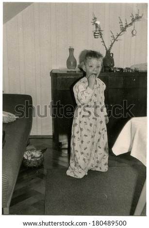 GERMANY - CIRCA 1960s: An antique photo of boy in a robe standing between table and sofa and eat candy behind him nightstand with spruce branch decorated with Christmas toys