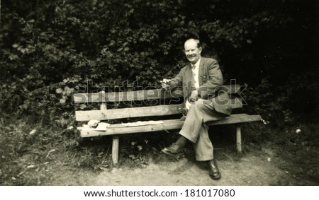 GERMANY - CIRCA 1950s: An antique photo of bald middle-aged man in a business suit with a cigar in hand posing on the garden bench