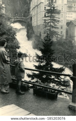 AUSTRIA, BAD GASTEIN - CIRCA 1958: An antique photo of woman and boy on the observation deck watching a torrent mountain Wrekin background multi-storey building