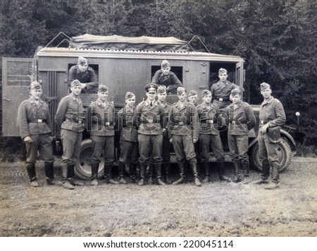 GERMANY - CIRCA 1940s: Group of German soldiers led by an officer posing against the backdrop of the truck facing the trees, Germany, 1940s. Reproduction of antique photo.