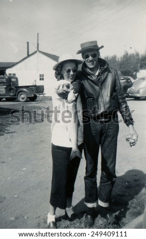 CANADA - CIRCA 1950s: Reproduction of an antique photo shows A man and a woman in a hat posing against the backdrop of the building and a pickup truck. They keep a pike in his hands.