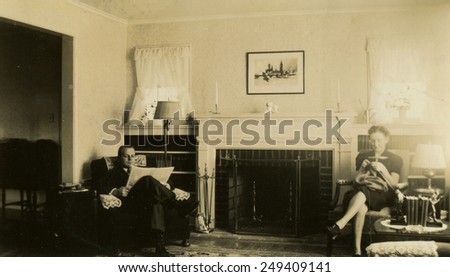 CANADA - CIRCA 1960s: Reproduction of an antique photo shows Man and woman sitting in a chair near the fireplace. A man reads a newspaper, woman knits on the needles.