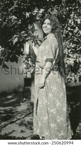 CANADA - CIRCA 1950s: Reproduction of an antique photo shows mature woman with the dismissed hair in a long dress posing with a smile on her face on the background of a flowering tree in the yard