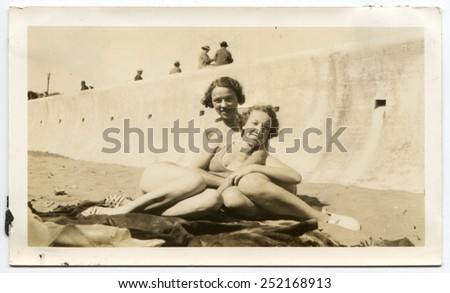 USA - CIRCA 1940s: Reproduction of an antique photo shows Two girls in bikini lying on a blanket on the sand on the beach, behind a concrete parapet are people