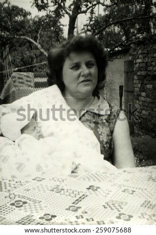 GERMANY - August 26, 1981: mature woman sitting in the garden near a table covered with a tablecloth, and holds a baby