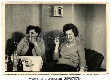 GERMANY - CIRCA 1960s: Two smiling women sitting at the banquet table in front of them is a bottle of vodka and two bottles of beer, a young woman smoking a cigarette