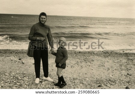 GERMANY - CIRCA 1950s: Woman in trousers and jumper, her head covered with a handkerchief, holding the hand of a little boy in shorts. They posing against the backdrop the sea.