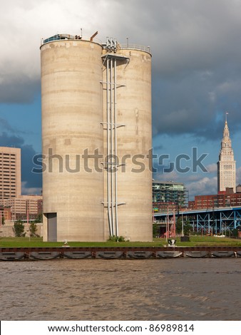 A pair of tall silos used to store powdered cement located on the bank of a river with a portion of the downtown city skyline and heavy clouds in the background