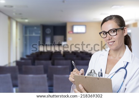 closeup of a friendly young female doctor with clipboard and a pen on a hospital waiting room background - focus on the doctor