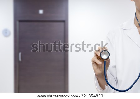 young female doctor holding with her right hand a stethoscope standing isolated on a medical consultation door background - focus on the hand