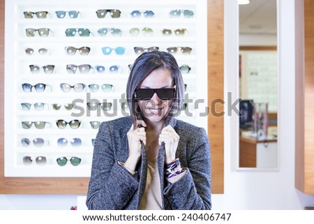 portrait of a young woman posing at the optical shop is trying on very fashionable sunglasses with the glasses expositor at the background - focus on the glasses center