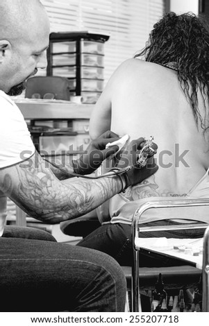 male tattoo artist is tattooing the back of a young woman in the tattoo cabin at his tattoo shop - focus on the tattoo machine