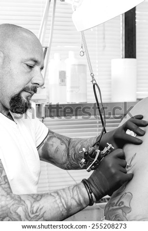 young tattooer is tattooing the back of a young woman in the tattoo cabin at his tattoo shop - focus on the man face