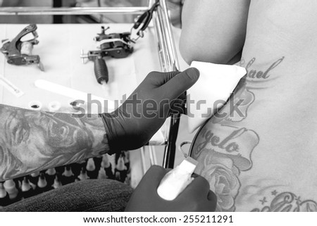 tattoo artist is spraying with a disinfectant the work area in a tattoo session at the tattoo shop - focus on the back of the woman