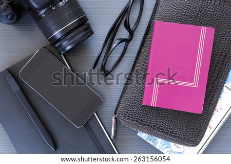 top view of a desktop with a travel stuff consisting on a camera, a passport, a documents case, a phone, a map, a reading glasses, a pen and a notebook on a desk background