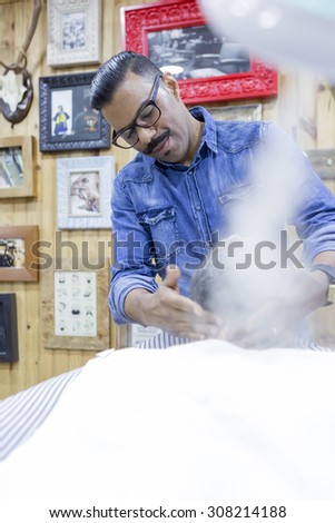 barber is steaming with a steam machine to a beard of a customer on a beard shaving session in a barber shop - focus on the left eye