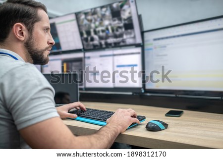 Monitoring. Attentive involved bearded young man with keyboard sitting watching in front of computer screens ストックフォト © 