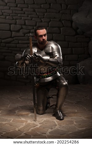 Medieval knight  kneeling with sword on a dark stone wall background