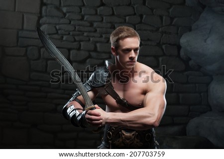 Half length portrait of young attractive warrior gladiator with muscular body posing with sword on dark background. Concept of masculine power, strength