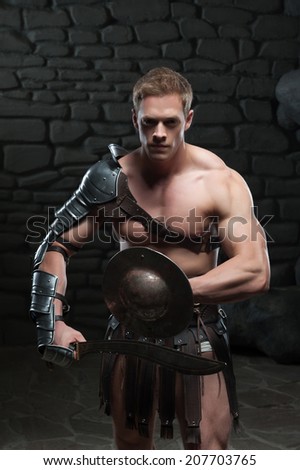 Half length portrait of young attractive warrior gladiator with muscular body posing with shield and sword on dark background. Concept of masculine power, strength.