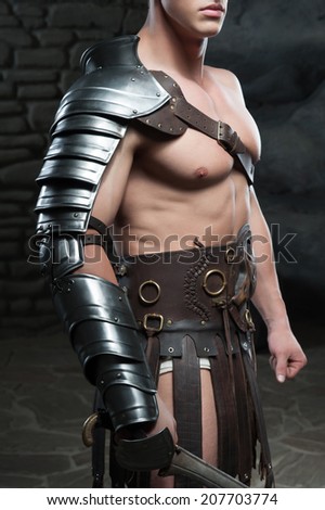 Closeup portrait of young attractive warrior gladiator with muscular body posing with sword on dark background. Concept of masculine power, strength