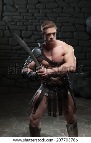 Half length portrait of young attractive warrior gladiator with muscular body posing with sword on dark background. Concept of masculine power, strength