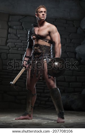 Full length portrait of young attractive warrior gladiator with muscular body holding shield and axe, posing on dark background. Concept of masculine power, strength