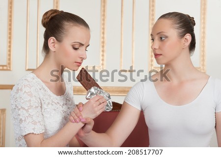 Two young beautiful ballet dancers posing with chocolate, one dancer keeps other from eating a bar of chocolate in classical interior. Concept of diet and wellness or keeping fit