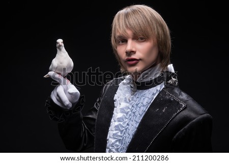 Half-length portrait of fair-haired matchless juggler wearing interesting black costume and white shirt holding pouter on his hand thinking about the next trick. Isolated on black background