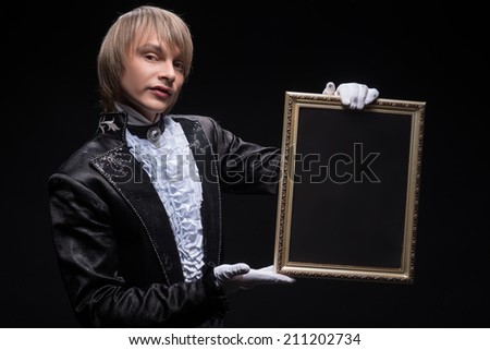 Half-length portrait of fair-haired matchless juggler wearing interesting black costume and white shirt showing us mysterious trick with the empty frame.Copy place. Isolated on black background