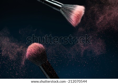 Horizontal close-up photo of  two professional  black make-up brushes with pink powder in motion isolated on blue background