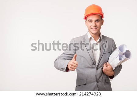 Half-length portrait of young smiling handsome architect engineer in orange helmet posing with blueprints showing thumb up isolated on white background, copyspace