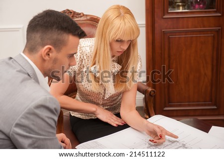 Top view portrait of young beautiful woman client discussing ground plan with handsome architect sitting at the table in design studio,  pointing at architectural plan, creative discussion concept
