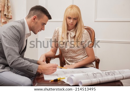 Close-up portrait of young handsome architect discussing ground plan with client sitting at the table in design studio,  pointing at architectural plan with pencil, creative discussion concept