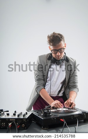 DJ playing music with stylish haircut and glasses and headphones with grey jacket at work spinning on mixer looking down while standing isolated on white background