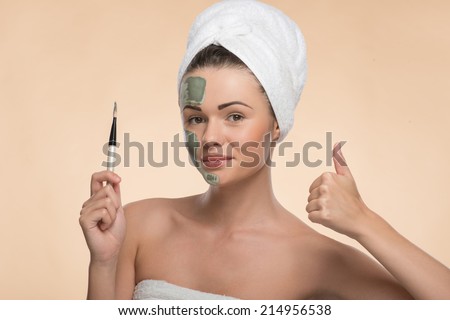 Horizontal portrait of happy smiling and looking at the camera girl in spa with  towel on her head applying facial clay mask with brush and showing sign OK isolated on beige background with copy place
