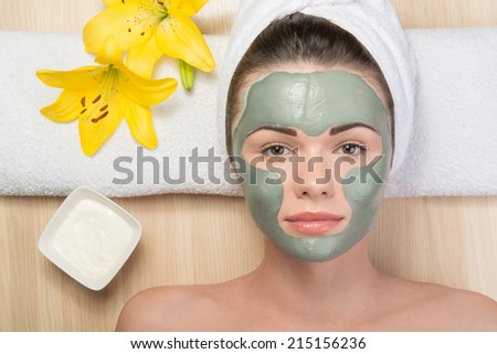 Close-up portrait of beautiful girl looking at the camera with a towel on her head applying facial clay mask and beauty treatments lying on a table in spa near yellow flower and white plate