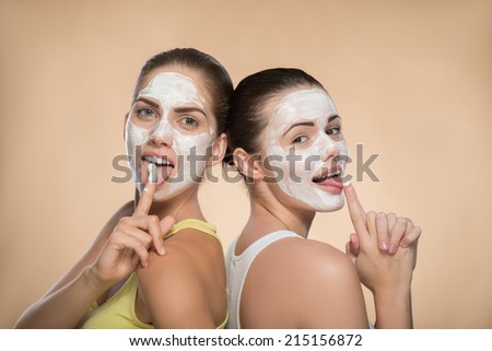 Portrait of two beautiful funny girls with cream on their faces standing back to back, one girls showing sign attention and other girl showing a gun isolated on beige background with copy place