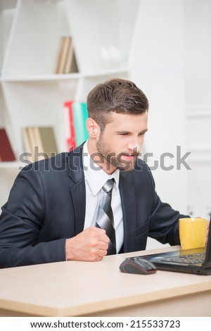 Portrait of handsome angry businessman sitting at the table in office having the coffee-break drinking yellow cup of tea and looking with irritation at his laptop