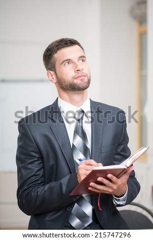 Waist-up portrait of handsome confident businessman sitting at the table and attentively writing some notes in red notebook in office interior and looking up