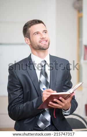 Waist-up portrait of handsome confident businessman sitting at the table and attentively writing some notes in red notebook in office interior and looking up and happy smiling