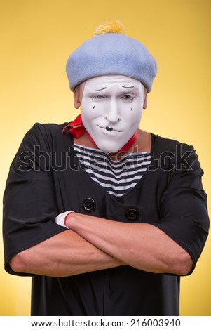 Waist-up portrait of pert mime with white face, grey hat and red scarf playfully looking at the camera folding his hands on his chest isolated on yellow background