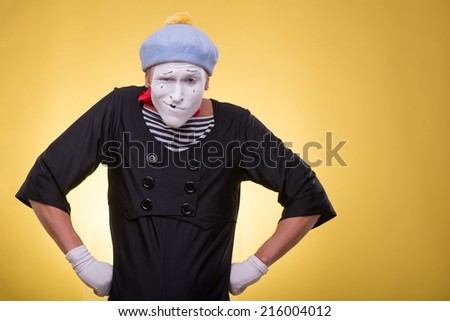 Waist-up portrait of pert mime with white face,  grey hat and red scarf playfully looking at the camera holding his hands on his hips isolated on yellow background