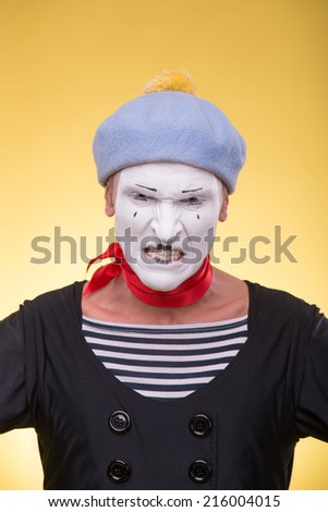 Waist-up portrait of angry mime with white face, grey hat and red scarf looking at the camera with irritation isolated on yellow background