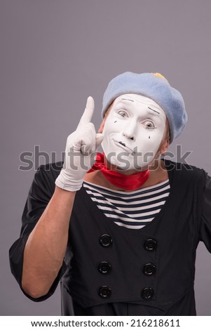 Waist-up portrait of funny male mime with grey hat and white face playfully looking at the camera and showing sign Attention isolated on grey background with copy place